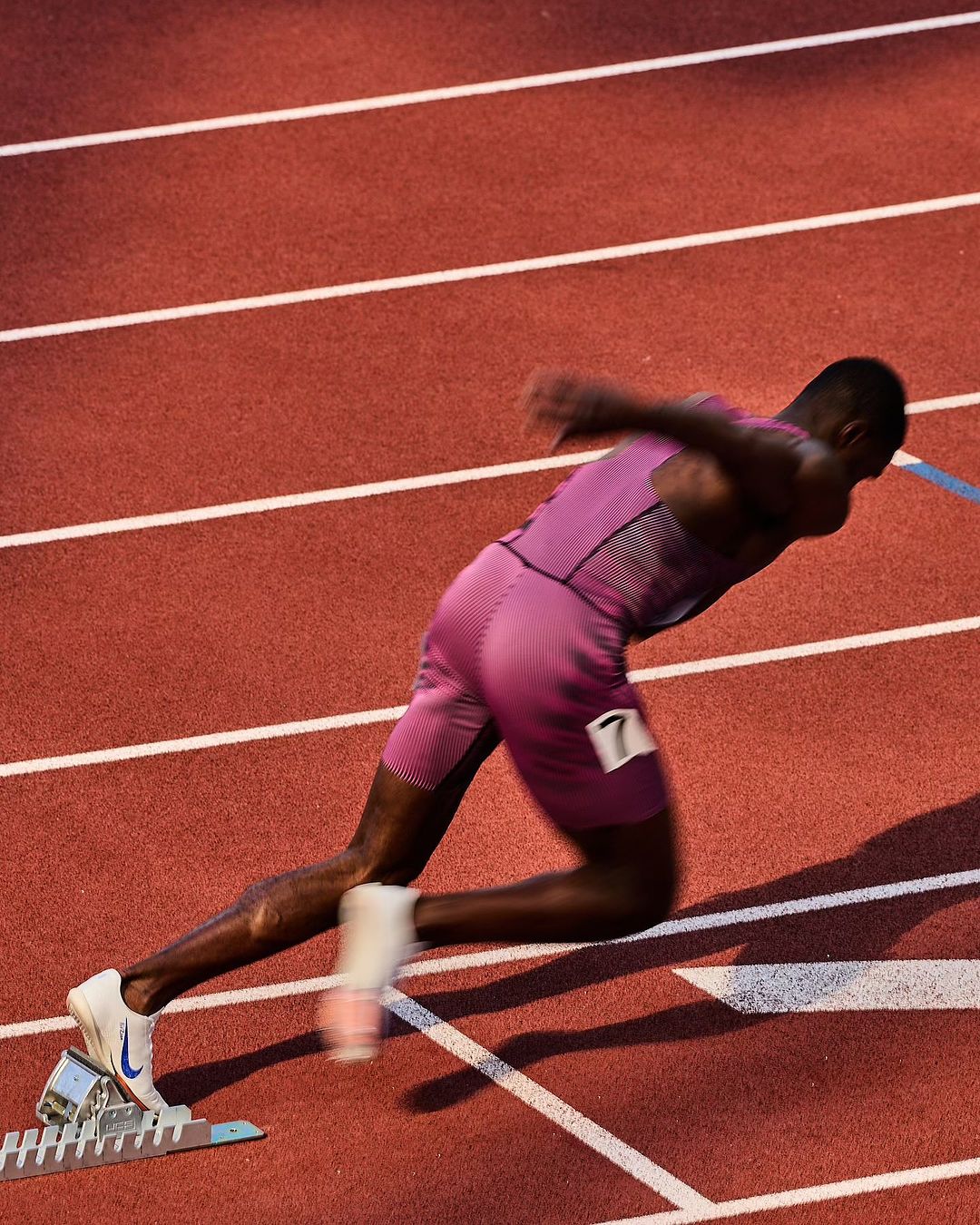 A sprinter in a pink uniform starts running from blocks on a red running track during a sports event at night under bright lights.