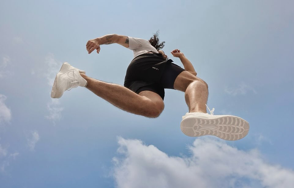 Low-angle shot of an adult man in a light t-shirt and black shorts jumping high against a blue sky with light white clouds.