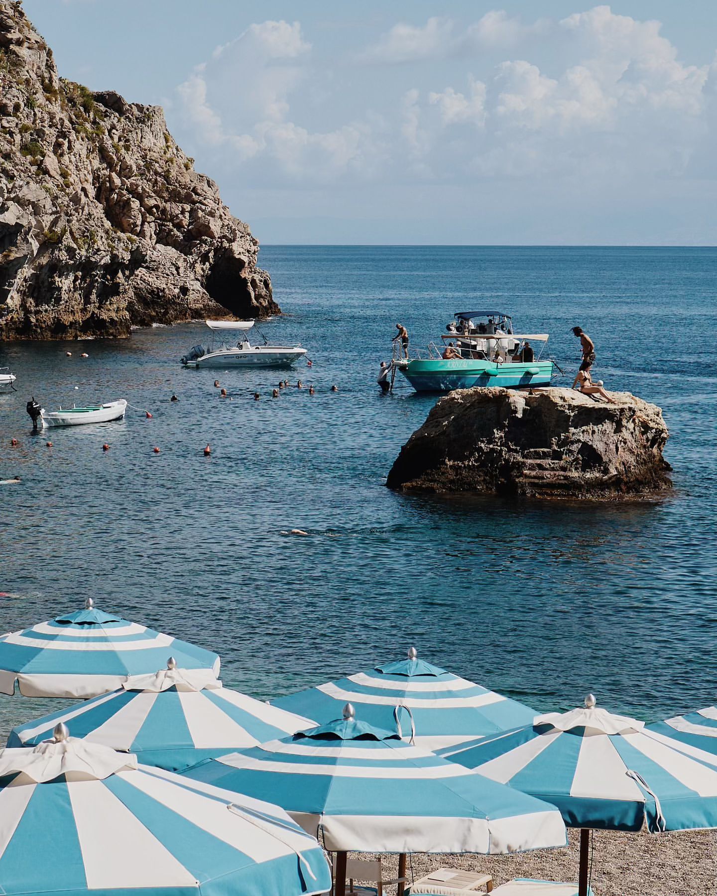 Scenic view of a beach with striped umbrellas, turquoise water, boats, and rocky cliffs under a sunny sky on the coast.