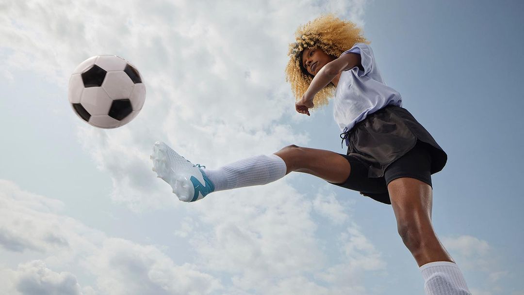 Woman kicks soccer ball mid-air in an upward, low-angle shot. She wears athletic clothing against a backdrop of cloudy sky, emphasizing motion and sport.