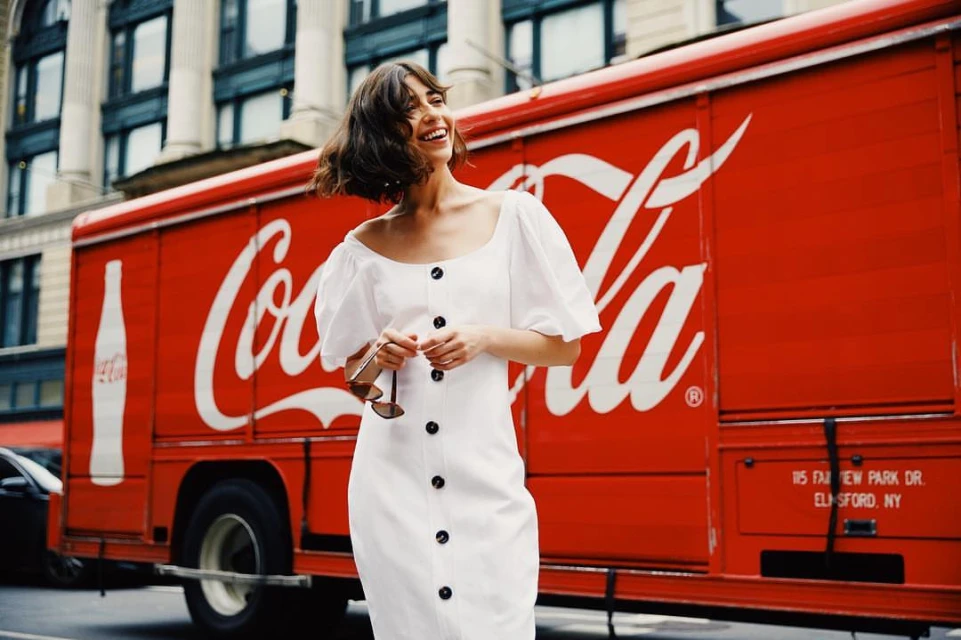 Woman in a white dress stands smiling in front of a vintage red Coca-Cola truck, posing on a city street with buildings in the background.