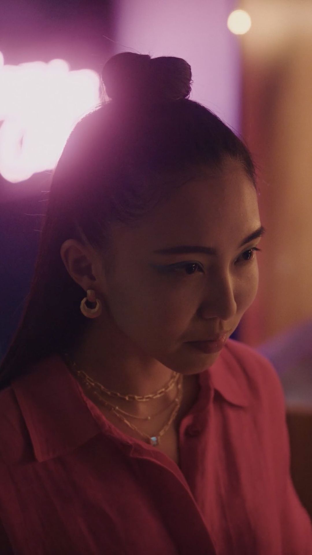 Close-up of an Asian woman with a pink shirt and gold necklaces, looking down, with a neon sign in the background.