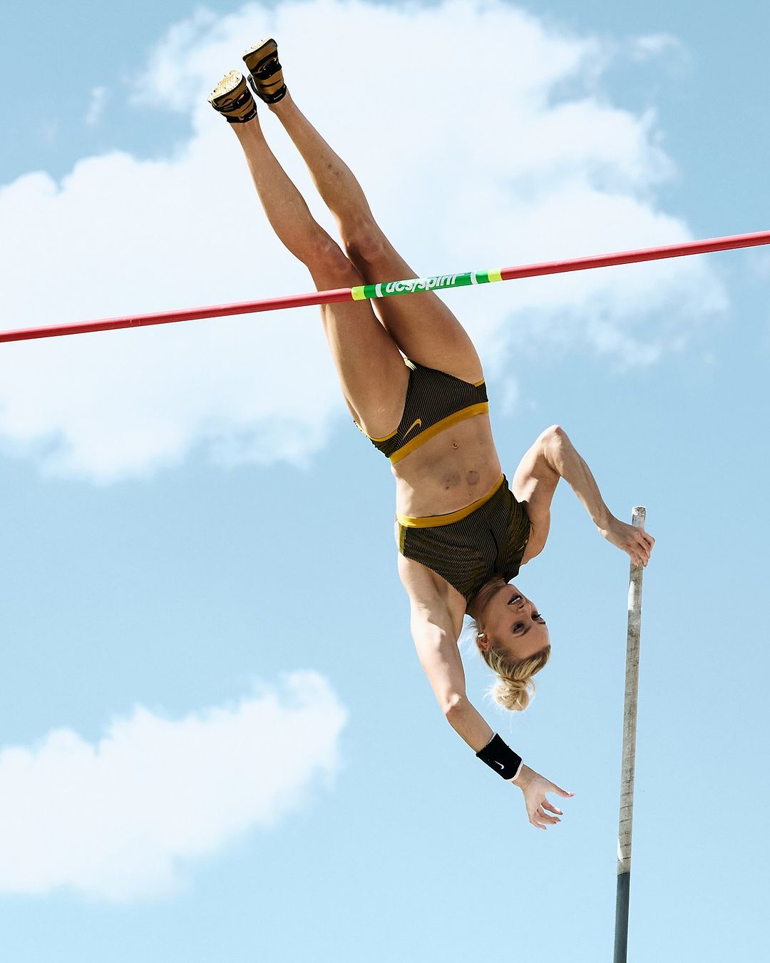 Female athlete in mid-air during a pole vault against a blue sky with white clouds. She's in athletic wear, exhibiting strength and skill.
