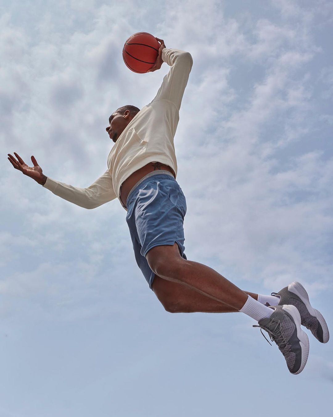 An athletic adult male leaps through the air with a basketball on an overcast day, wearing casual athletic wear with shoes and socks.