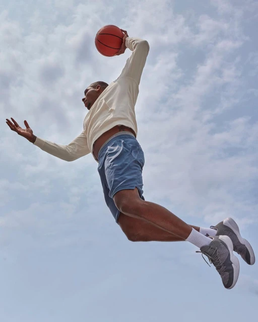 An athletic adult male leaps through the air with a basketball on an overcast day, wearing casual athletic wear with shoes and socks.