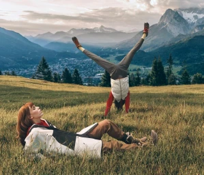 Two people in a grassy alpine meadow with mountain peaks in the background, one lying down and one doing a handstand.