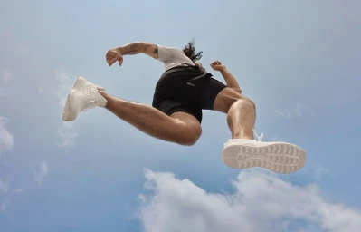 Low angle shot of a man leaping in the air wearing white sneakers against a pale blue sky with scattered clouds.
