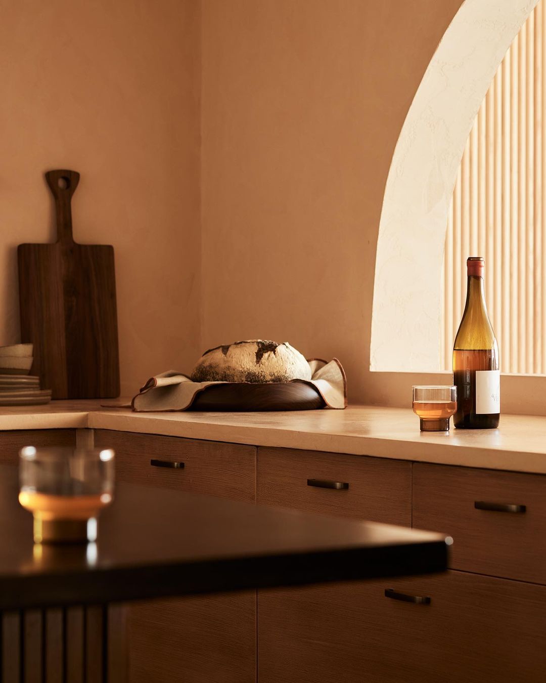 Still life shot of freshly baked bread on a wooden plate, a bottle of wine, and a glass in a warm-toned kitchen setting.