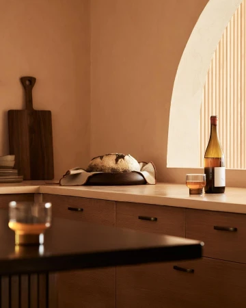 Still life shot of freshly baked bread on a wooden plate, a bottle of wine, and a glass in a warm-toned kitchen setting.