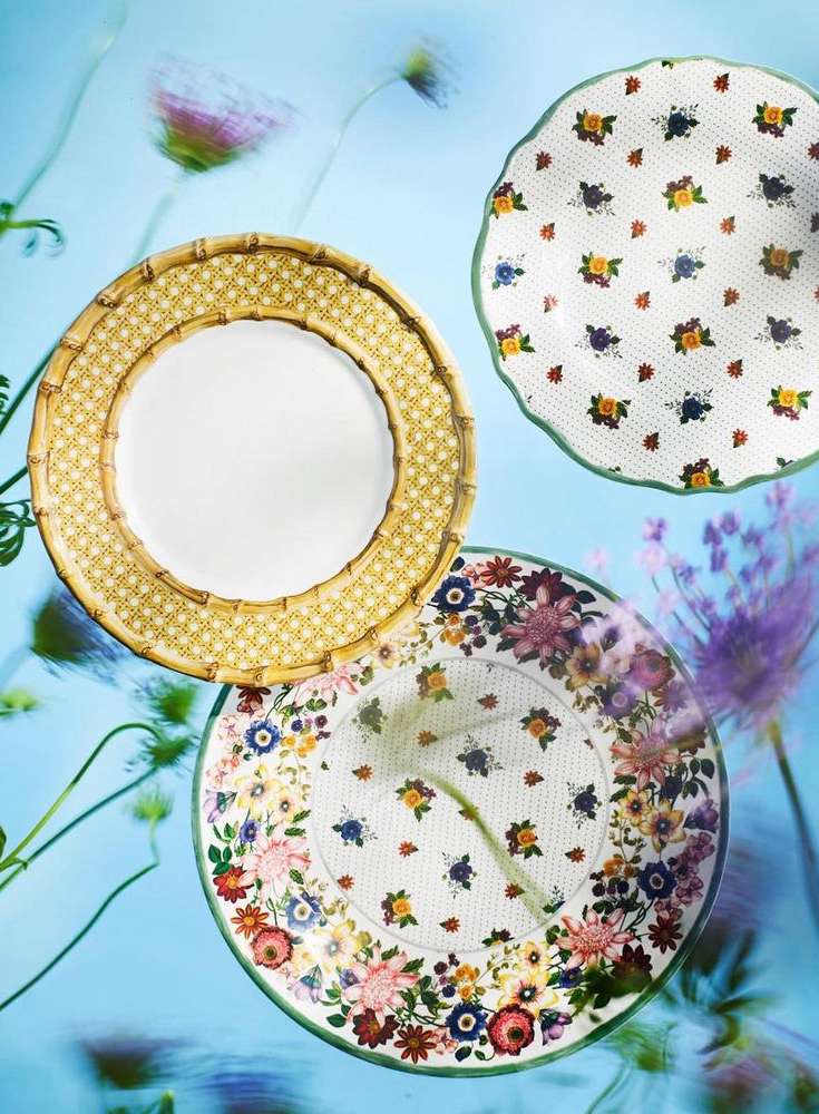 Overhead shot of three floral-patterned plates arranged on a blue background, soft focus floral accents, dinnerware composition.