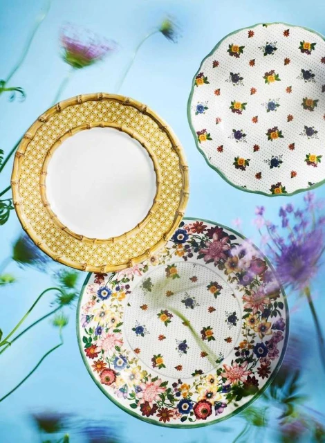 Overhead shot of three floral-patterned plates arranged on a blue background, soft focus floral accents, dinnerware composition.