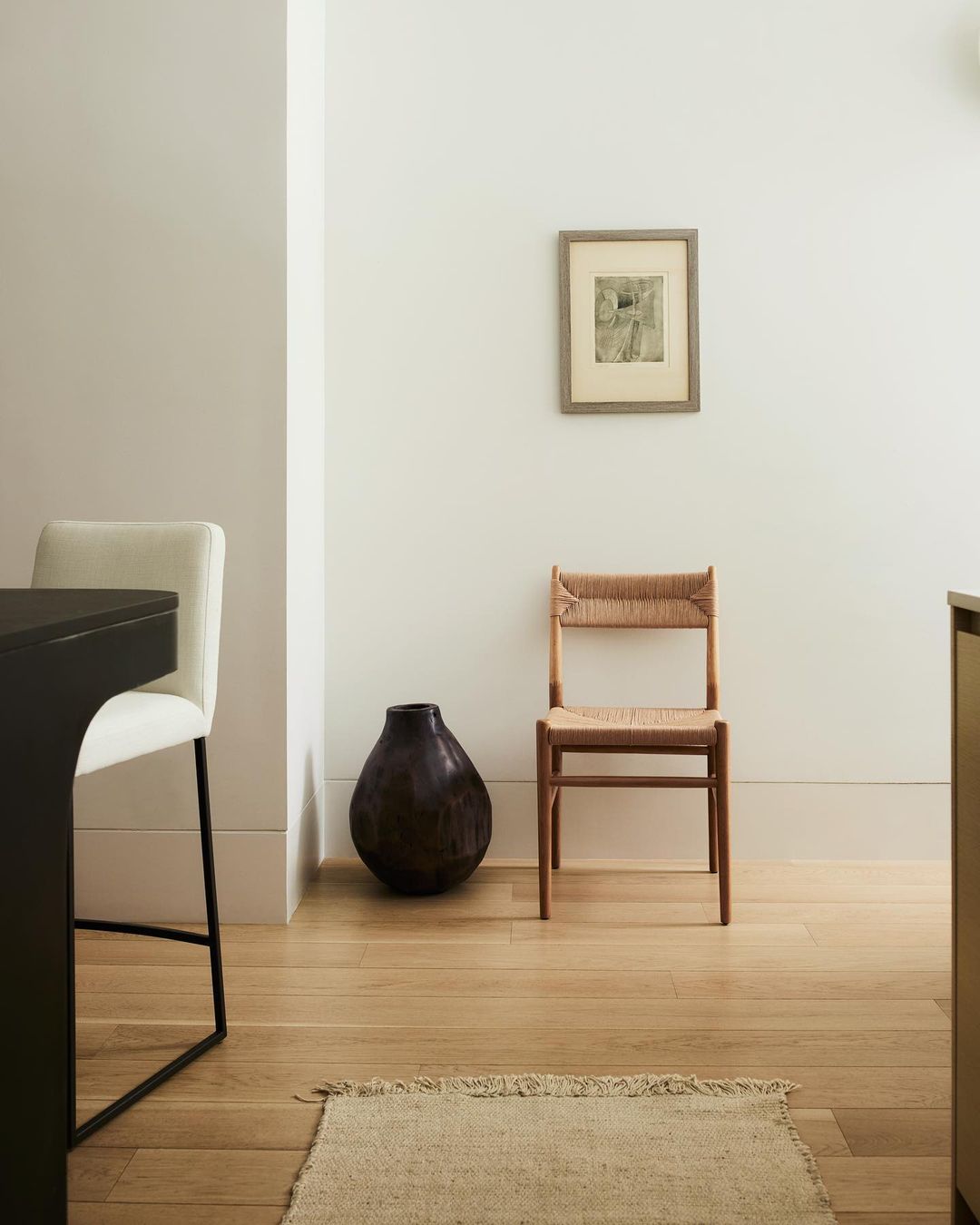 Interior design with a wooden chair, dark vase, framed art, and a neutral rug on light wood floors against white walls.