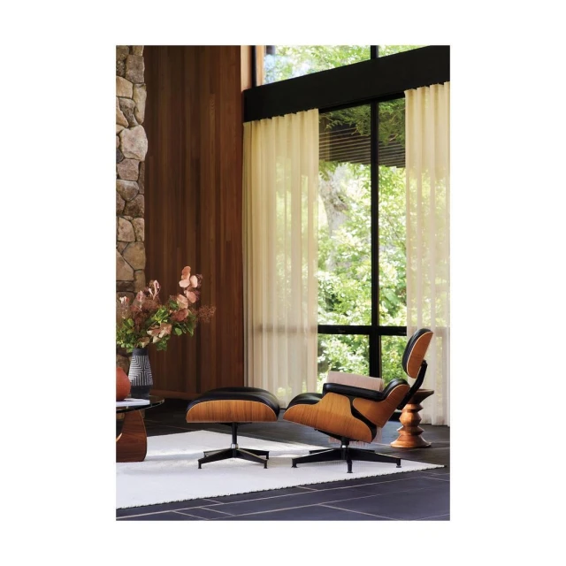 Eames lounge chair and ottoman in a modern living room with a stone fireplace, wood paneling, and sheer curtains.