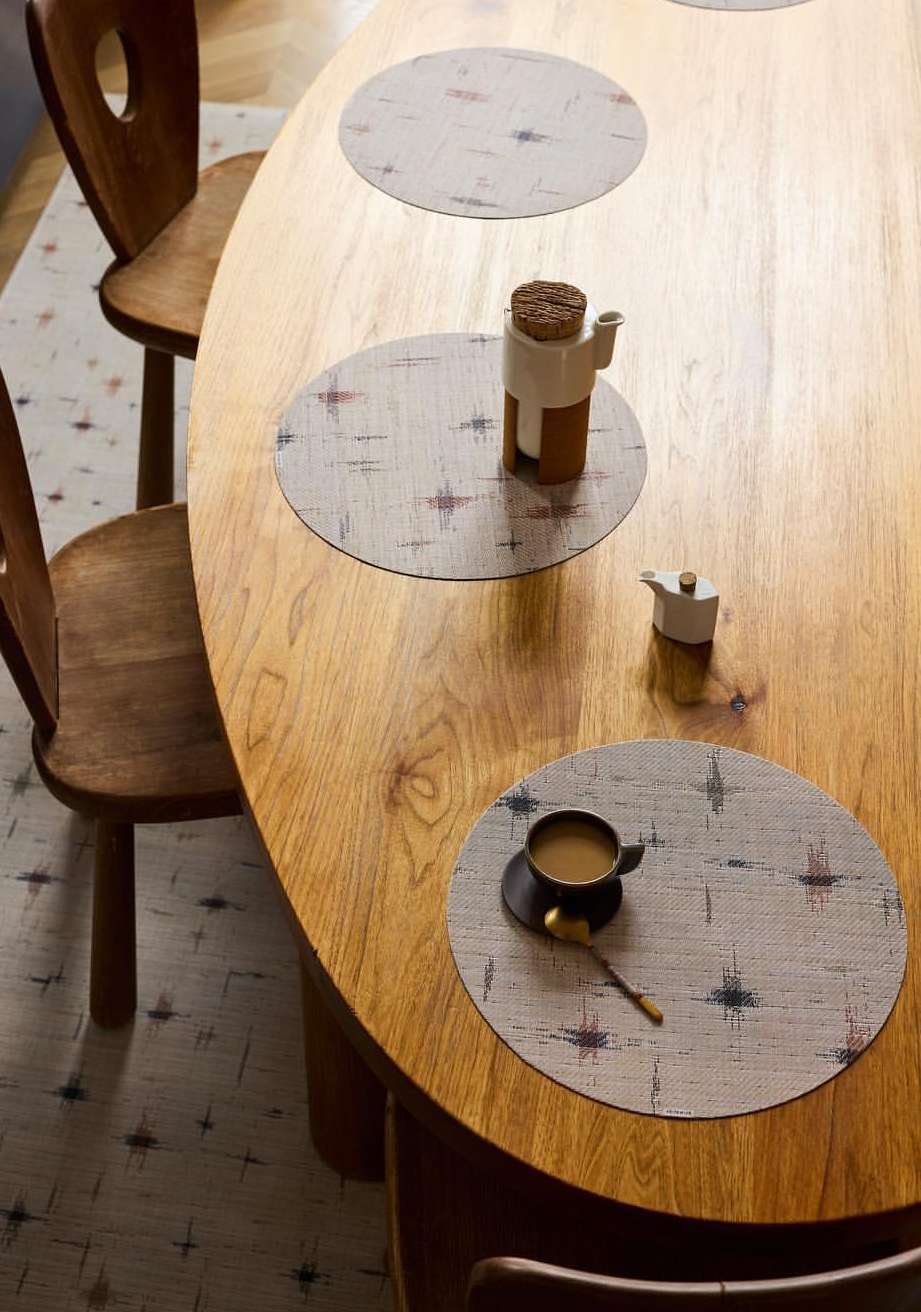Overhead view of wooden table set with neutral-toned patterned placemats, teapot, coffee cup, and wooden chairs, suggesting a warm, inviting atmosphere.