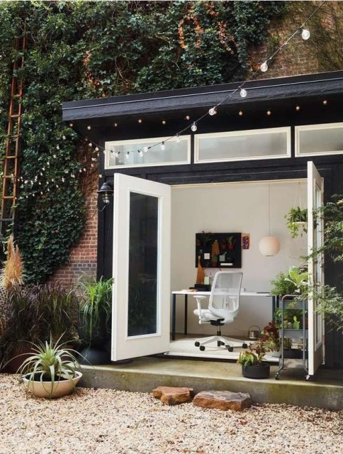 Outdoor office shed with open white doors, plants, gravel ground, string lights, and a modern white desk and chair visible inside.