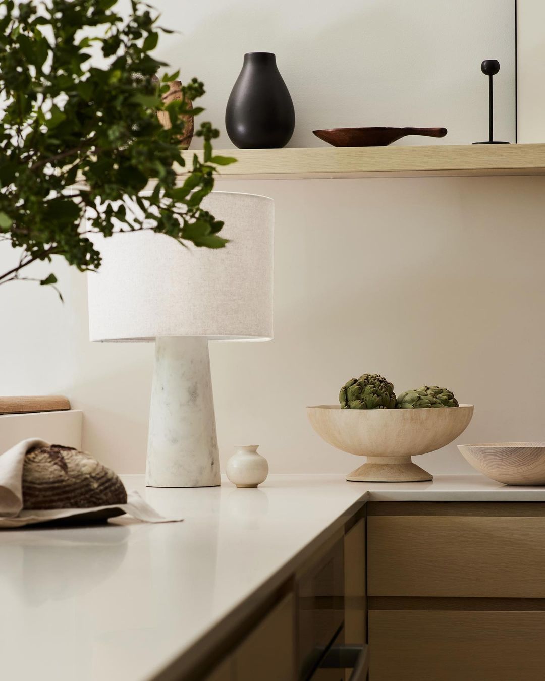 Still life composition featuring a lamp, wooden bowl with artichokes, a vase, and bread on a white countertop in a kitchen setting.
