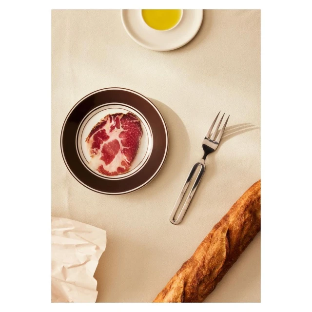 Overhead shot of charcuterie on a brown-rimmed plate with a fork, olive oil, bread, and crumpled paper on a beige surface.