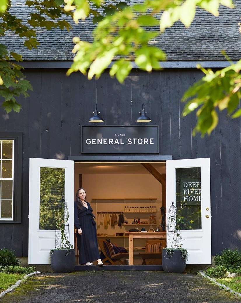 Exterior of a general store with a woman standing in the open doorway, surrounded by lush greenery and natural light.