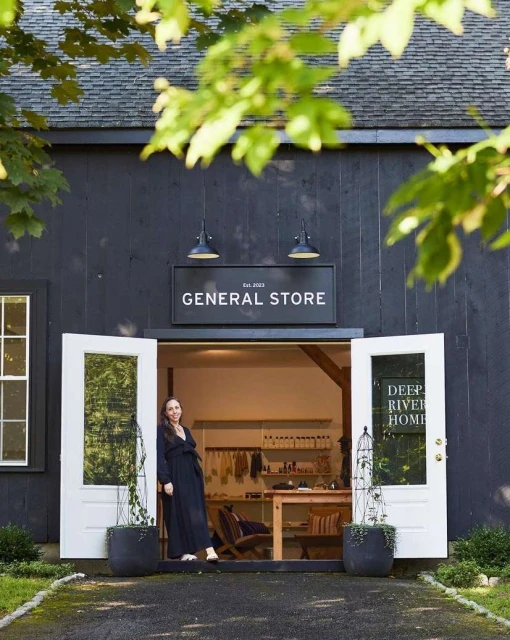Exterior of a general store with a woman standing in the open doorway, surrounded by lush greenery and natural light.