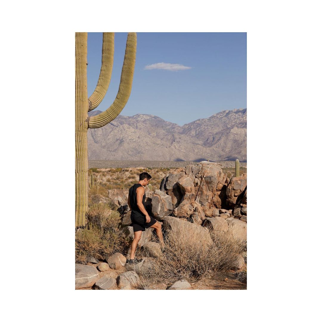 Athletic man in workout wear walks through desert landscape with cactus, rock formations, shrubs, and distant mountains under blue sky.