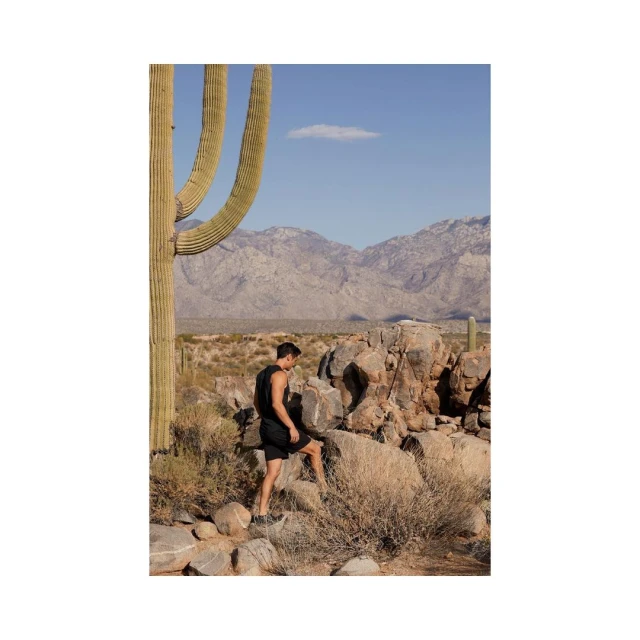Athletic man in workout wear walks through desert landscape with cactus, rock formations, shrubs, and distant mountains under blue sky.