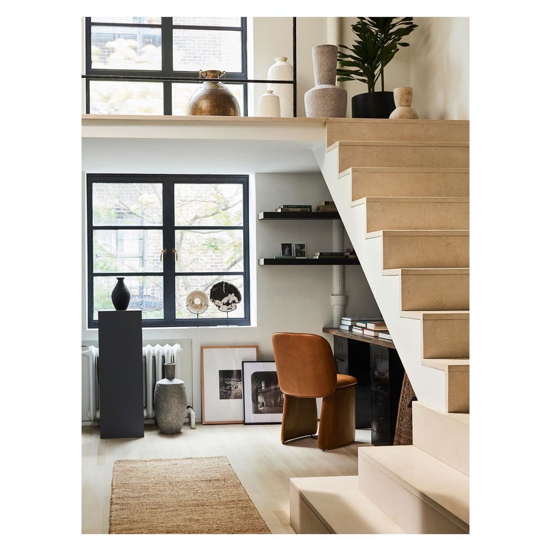 Stylish home interior with vases, stairs, and natural light through windows. Jute rug on hardwood floor and brown chair near a desk.