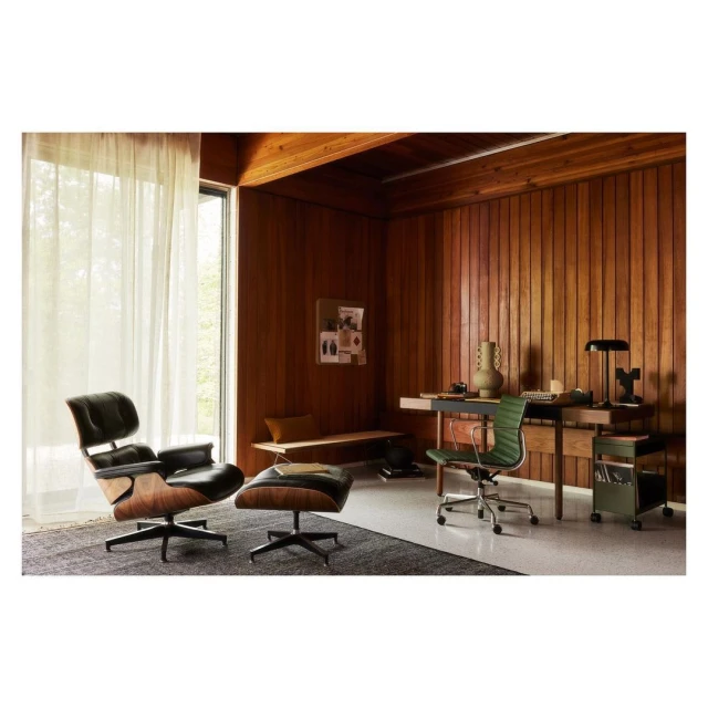 Interior view of a mid-century modern styled room featuring a black leather lounge chair and footstool, a wooden desk, and a rolling cart.