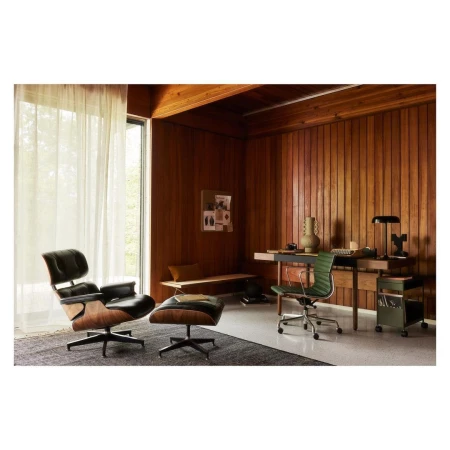 Interior view of a mid-century modern styled room featuring a black leather lounge chair and footstool, a wooden desk, and a rolling cart.