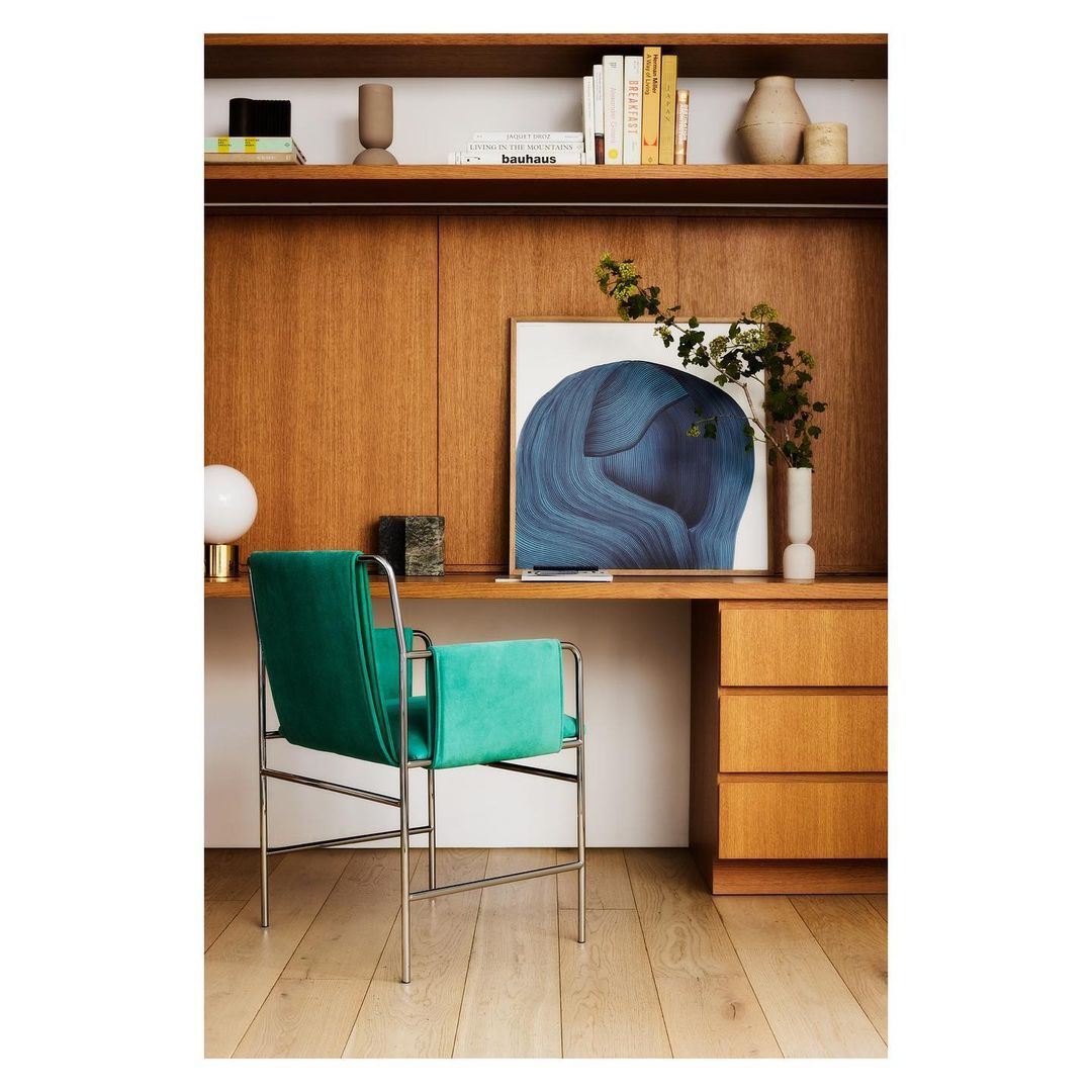 Interior design featuring a green chair, wood desk, and abstract art in a well-lit room. Books and decorative objects on a shelf.