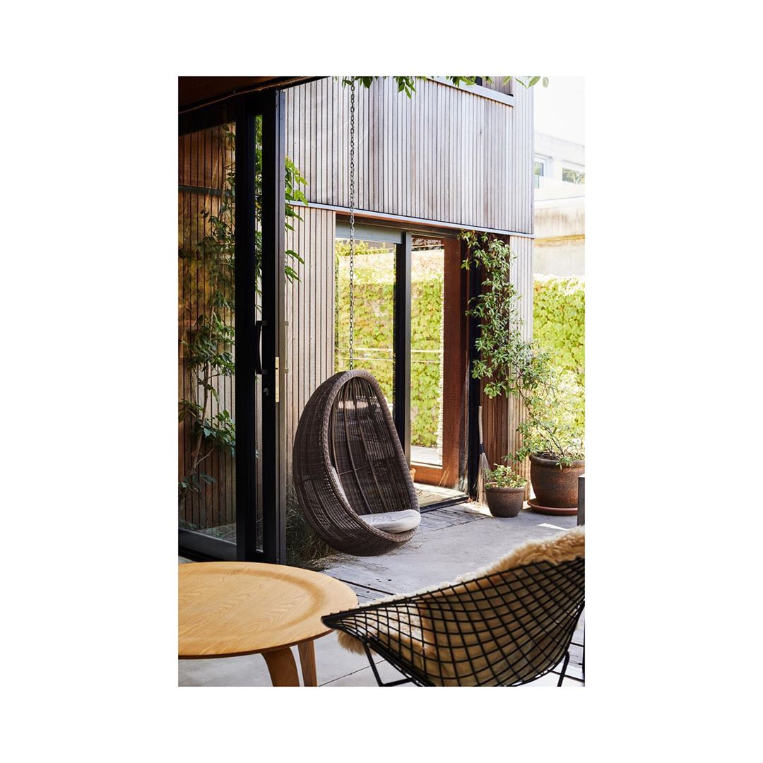 Outdoor patio area with a hanging egg chair, modern chair, small table, potted plants, and a wooden exterior wall on a sunny day.