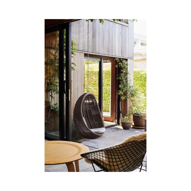 Outdoor patio area with a hanging egg chair, modern chair, small table, potted plants, and a wooden exterior wall on a sunny day.