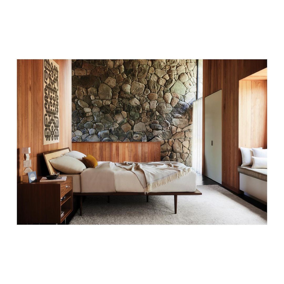 Bedroom interior with bed, stone wall, wood panelling, carpet floor, dark wood nightstand, and patterned textile art on the wall.