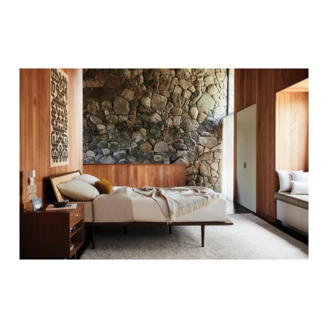 Bedroom interior with bed, stone wall, wood panelling, carpet floor, dark wood nightstand, and patterned textile art on the wall.