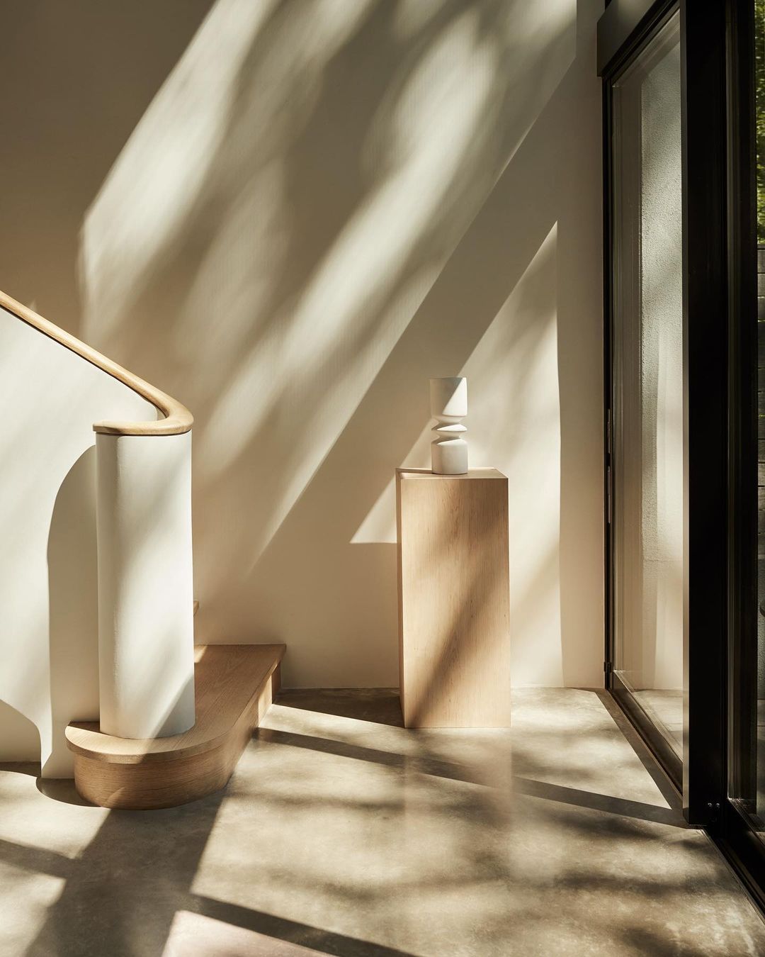 Modern interior featuring a white staircase, neutral wall, pedestal with a vase, and natural light streaming through a large window.