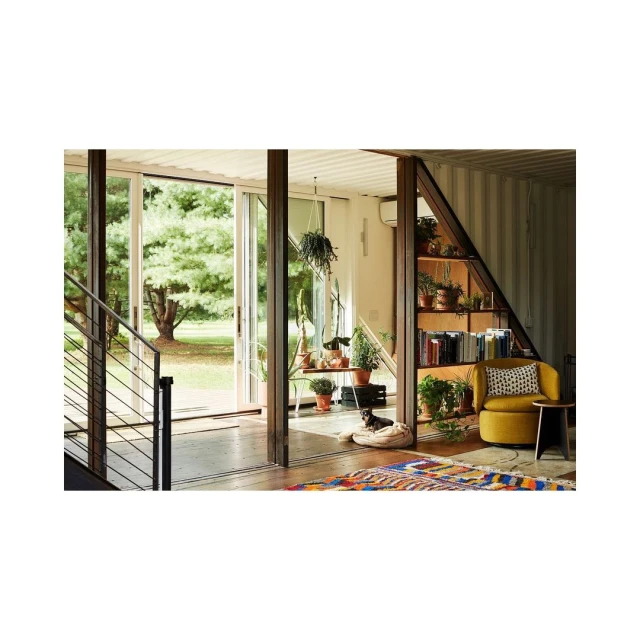 A living room interior featuring a dog in a dog bed, a yellow chair, a bookshelf, potted plants, and an outdoor view through glass doors.