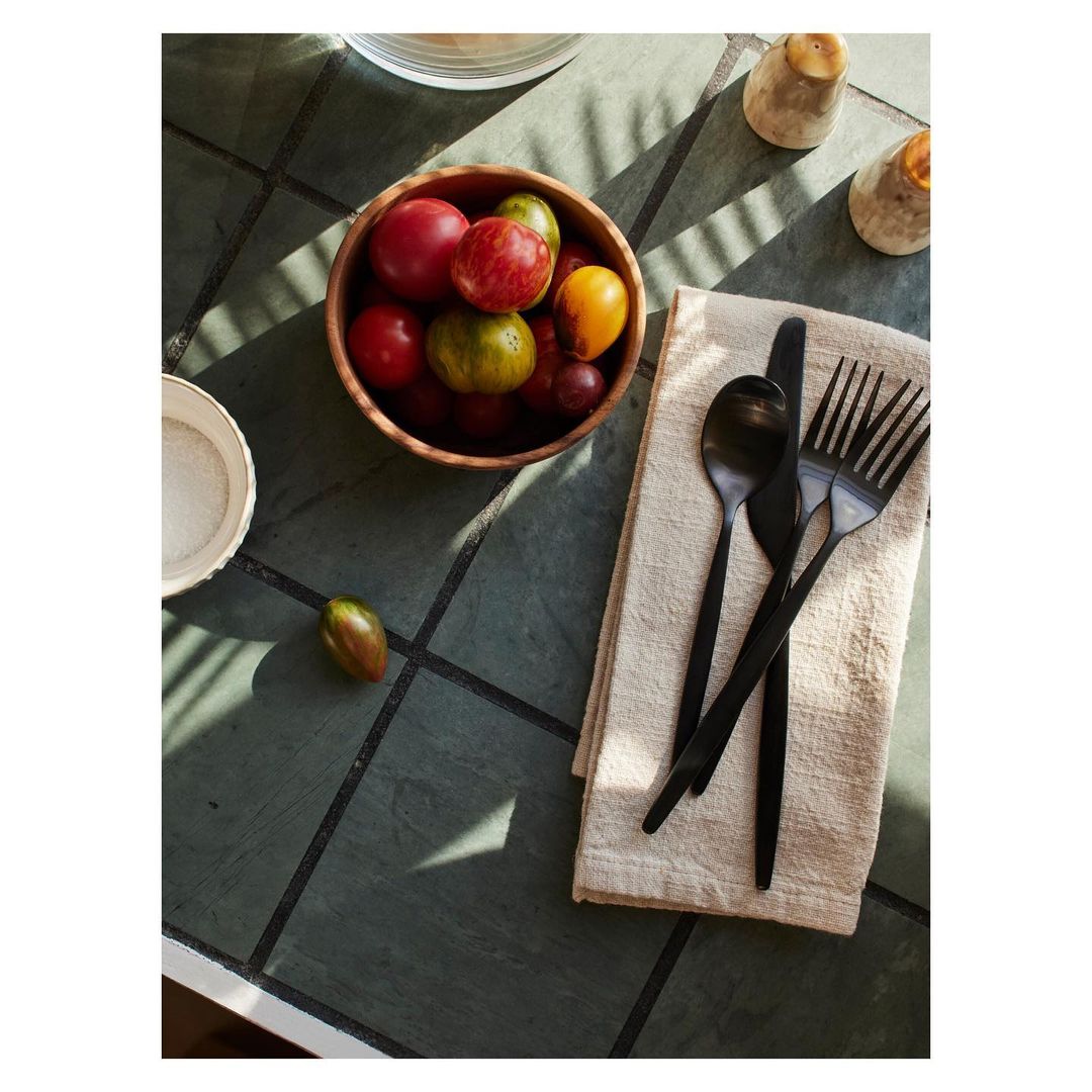 Overhead shot of heirloom tomatoes in wooden bowl, black flatware on beige linen napkin, and salt shakers on grey tiled table.