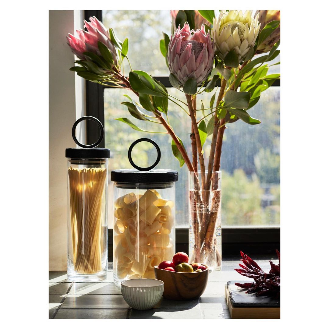 Still life arrangement features glass storage jars filled with pasta, accompanied by protea flowers in a glass vase against a window.