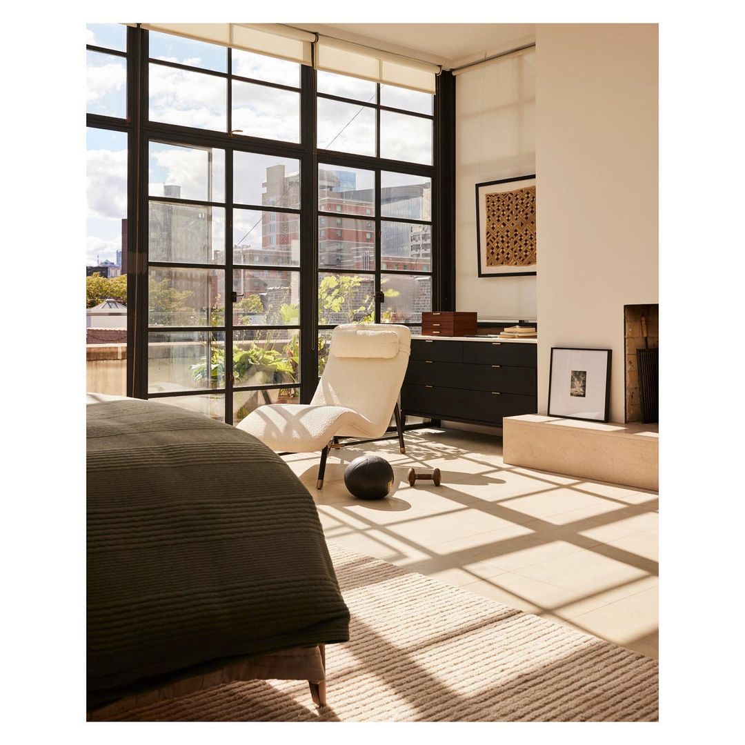 Modern bedroom interior featuring a chaise lounge by large windows with city views, complemented by neutral tones, and minimal decor.