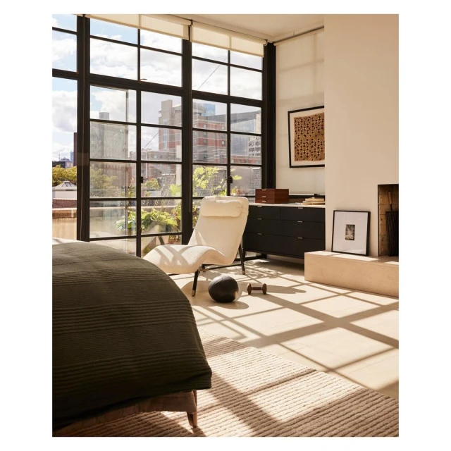 Modern bedroom interior featuring a chaise lounge by large windows with city views, complemented by neutral tones, and minimal decor.