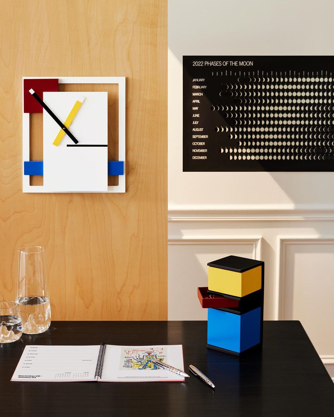Modern interior with a Mondrian-inspired wall clock, moon phase calendar, desk organizer, desk, and water glasses creating a neat composition.