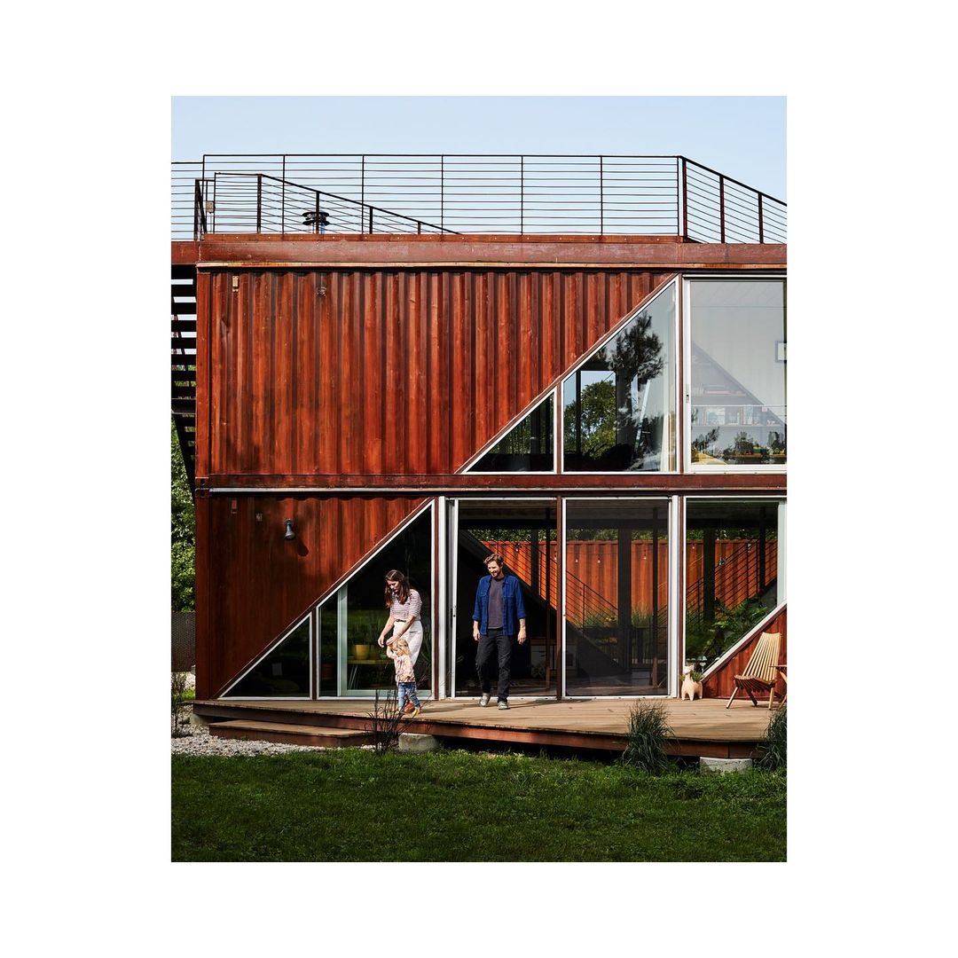 Two people standing on a wooden porch in front of a modern, two-story house built from shipping containers and large glass windows.