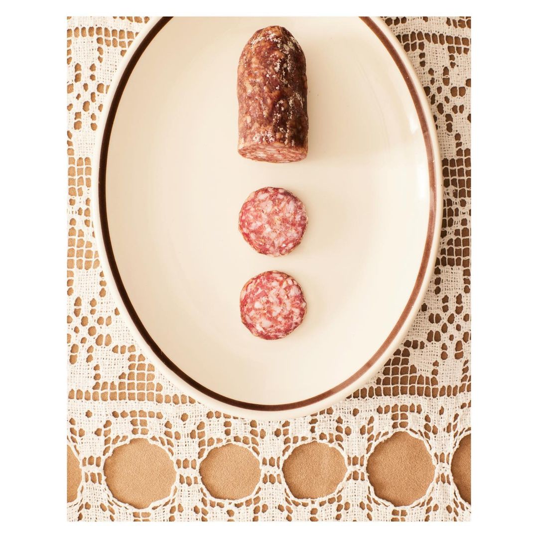 Overhead shot of a white plate with a piece of salami and two salami slices on a lace tablecloth. Food still life photography.