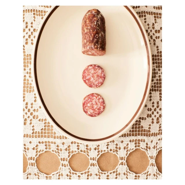 Overhead shot of a white plate with a piece of salami and two salami slices on a lace tablecloth. Food still life photography.
