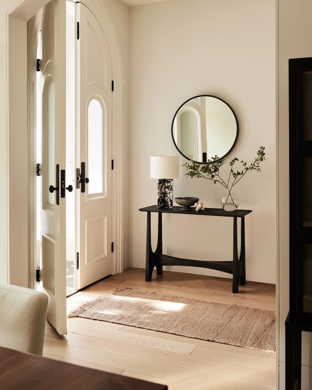 Interior entryway featuring white doors, a black console table with a lamp, and a round mirror against a light neutral wall.