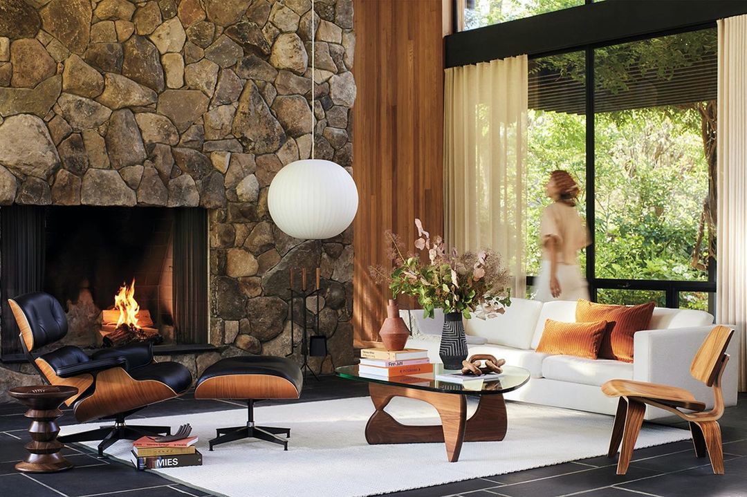 Living room interior with a stone fireplace, modern furniture including a white sofa, a wooden chair, and a hanging round light.