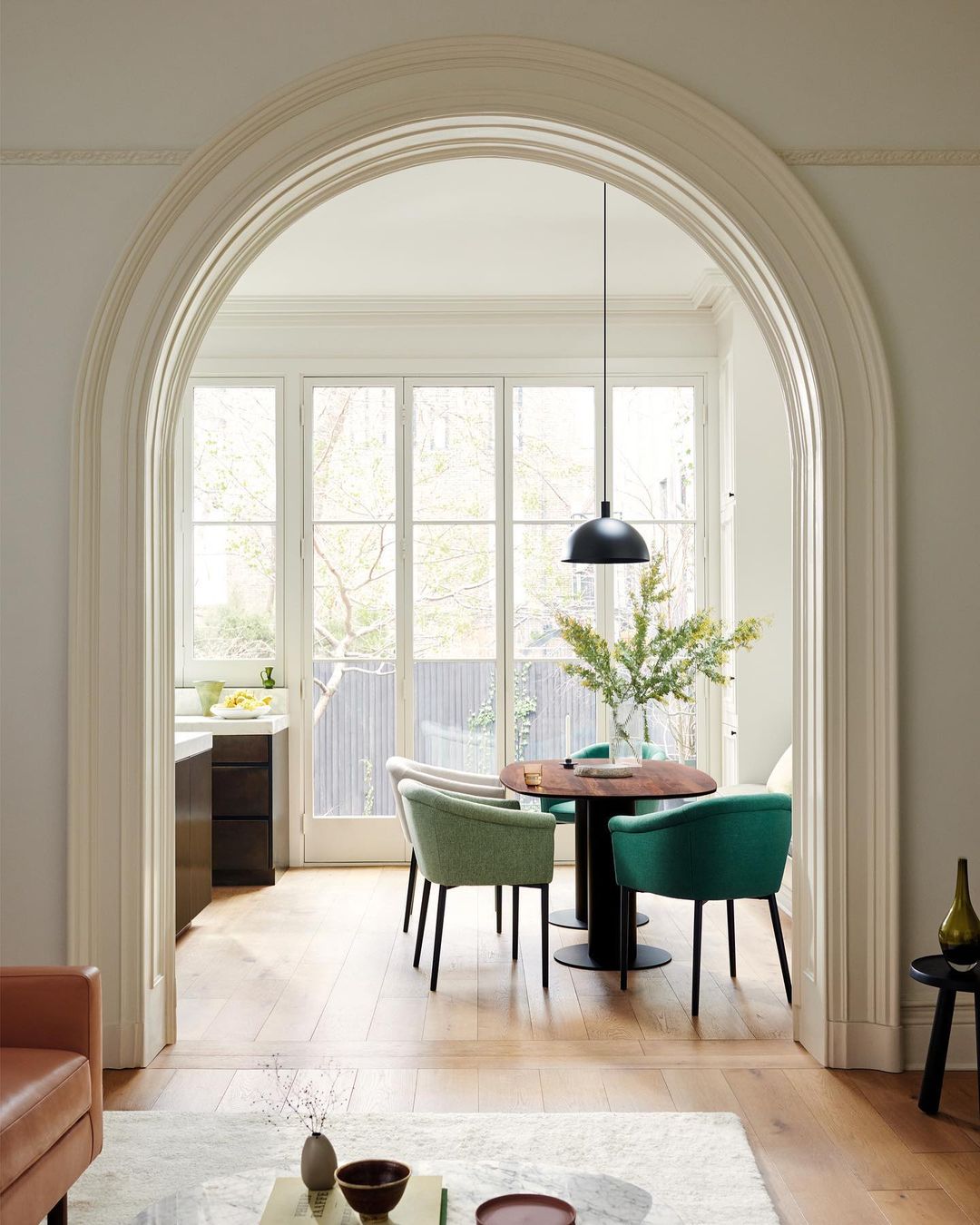 Interior view through arched doorway shows dining table with four chairs under pendant light, adjacent to sunlit window and kitchen area.