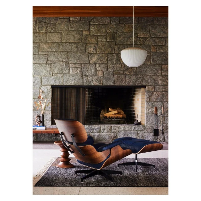 Eames lounge chair and ottoman with stone fireplace in interior setting. Decorative lighting and textured rug complete the interior.