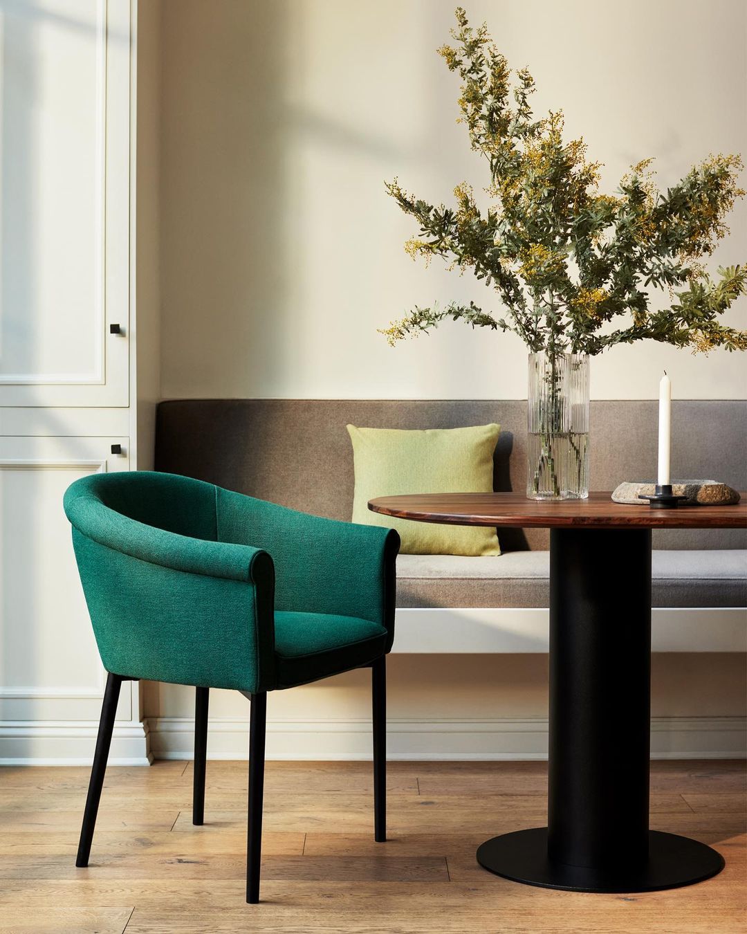 Green upholstered chair with black legs, round wood dining table with black pedestal base, floral arrangement, in neutral-toned dining space.