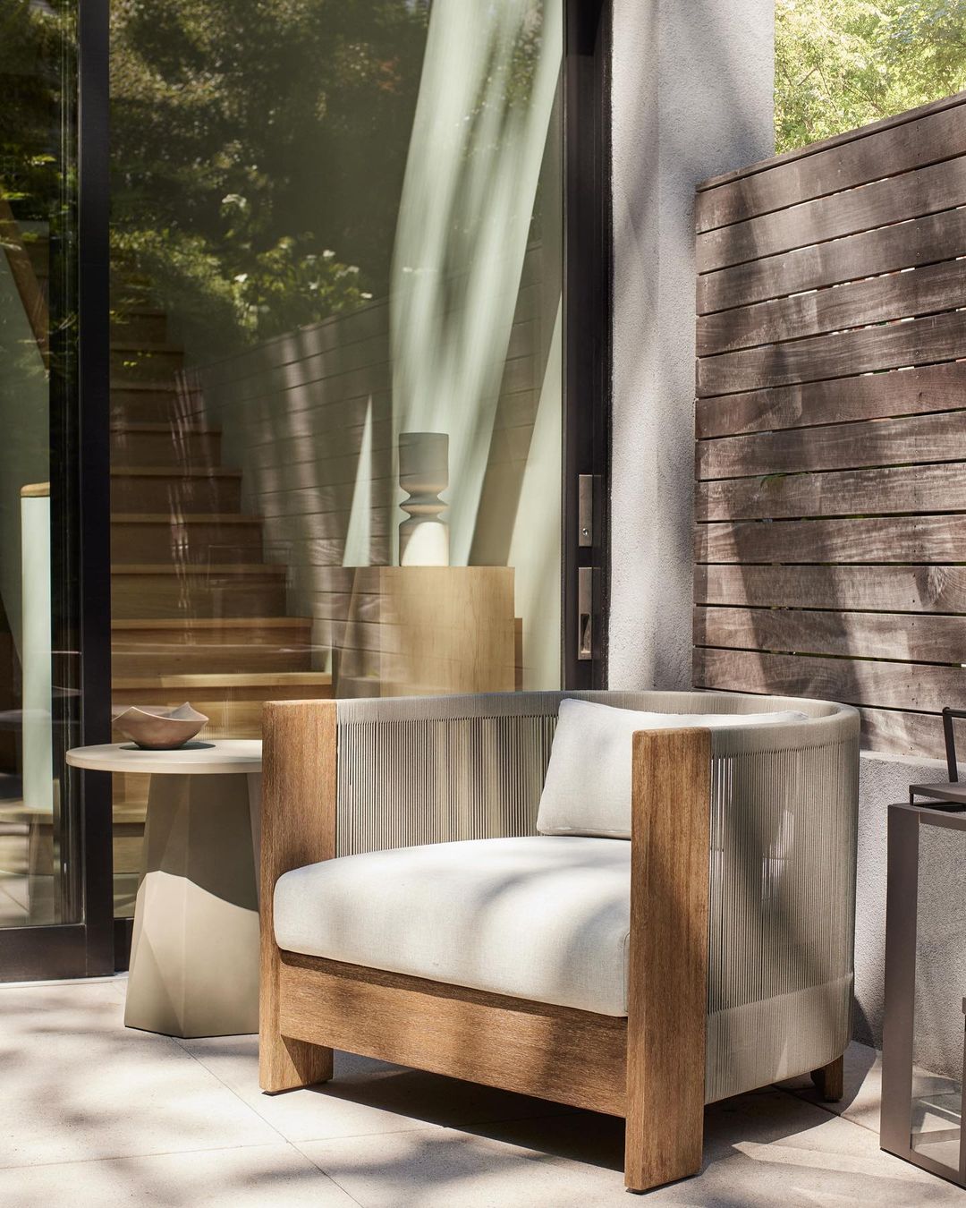 Patio seating with a modern wood frame chair and textured cushion, complemented by a side table and outdoor decor, set against a wooden fence.