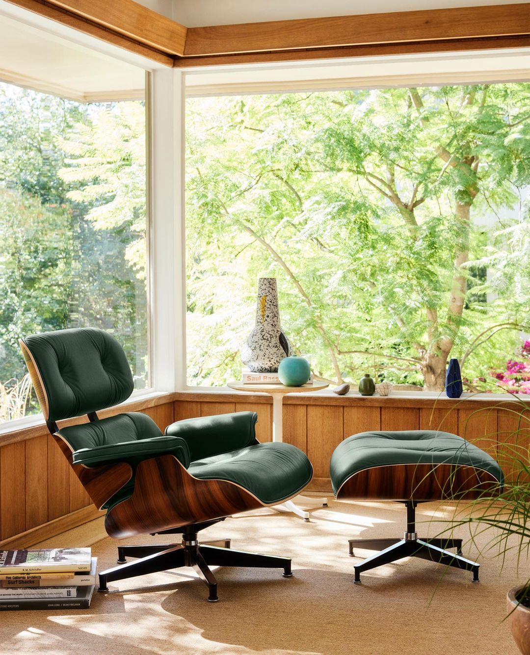 Eames lounge chair and ottoman in dark green leather and wood, with large window and leafy trees in background, in a bright room.